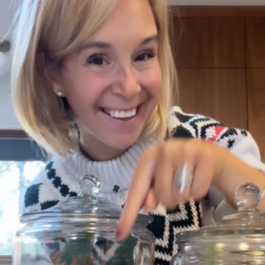 Smiling woman with short blonde hair, wearing a festive sweater, points at a glass jar containing small reindeer and a mini tree. Another jar with a mini tree is nearby. Warm indoor setting visible in the background.